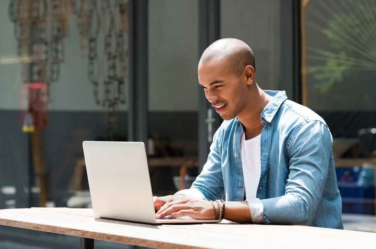 A man with short hair and glasses, dressed in a business-casual shirt, sits at a desk with a laptop in front of him. He appears focused and engaged as he works on the computer, which displays a digital marketing dashboard or course interface. The scene represents an AI-based premium digital marketing course setting.